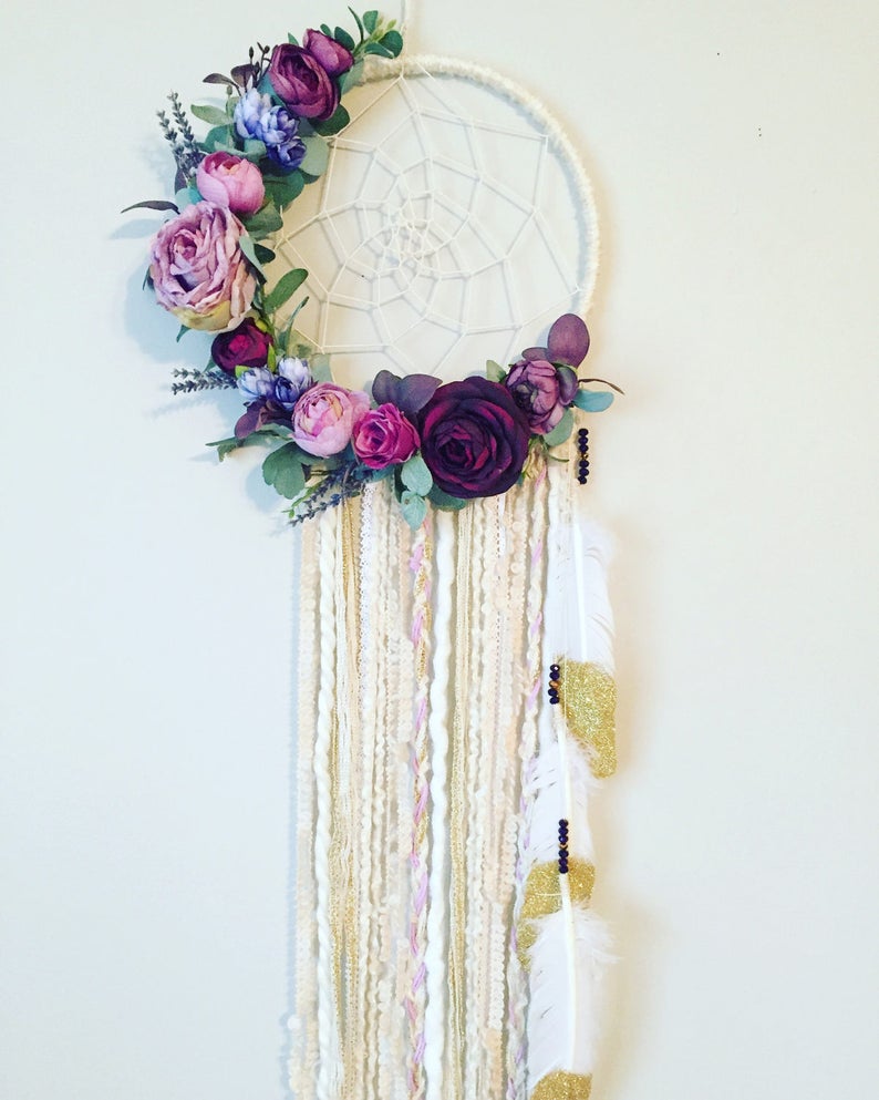 May include: A white dreamcatcher with a purple and pink floral wreath, white, gold, and pink yarn strands, and white feathers with gold glitter accents.