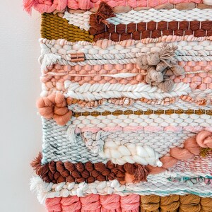 May include: A woven wall hanging with a pink, brown, white, and gold color scheme. The hanging features a variety of textures and patterns, including tassels and pom-poms.