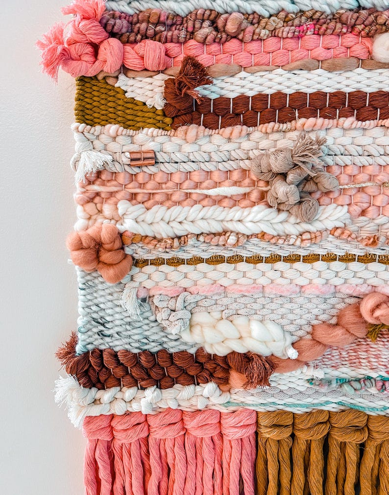 May include: A woven wall hanging with a pink, brown, white, and gold color scheme. The hanging features a variety of textures and patterns, including tassels and pom-poms.