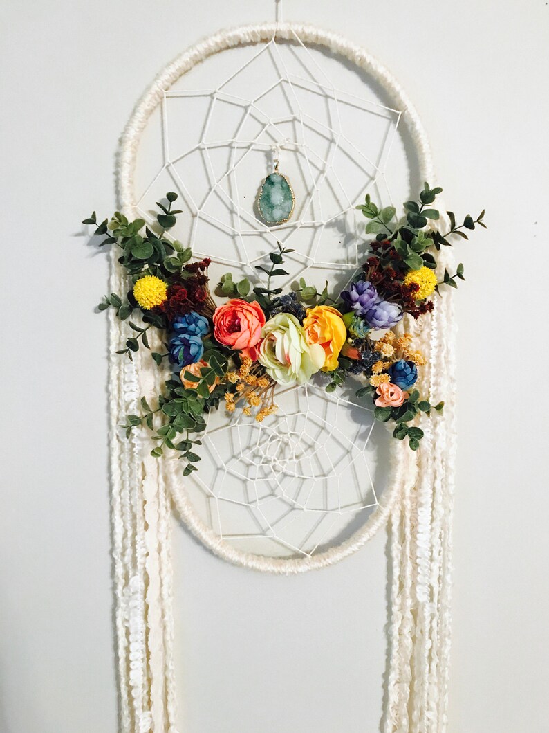 Puede incluir: Un atrapasue&ntilde;os blanco con una red de macram&eacute; y un colgante de piedra preciosa turquesa. El atrapasue&ntilde;os est&aacute; decorado con un arreglo floral de flores blancas, amarillas, rosas, azules y moradas.