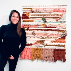 May include: A colorful woven wall hanging with a variety of textures and colors, including pink, orange, brown, and white. The hanging has a fringe border and is made with yarn.