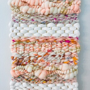 May include: A pink, white, and gold woven wall hanging with a textured, fluffy design. The hanging is made with yarn and has a variety of colors and textures.