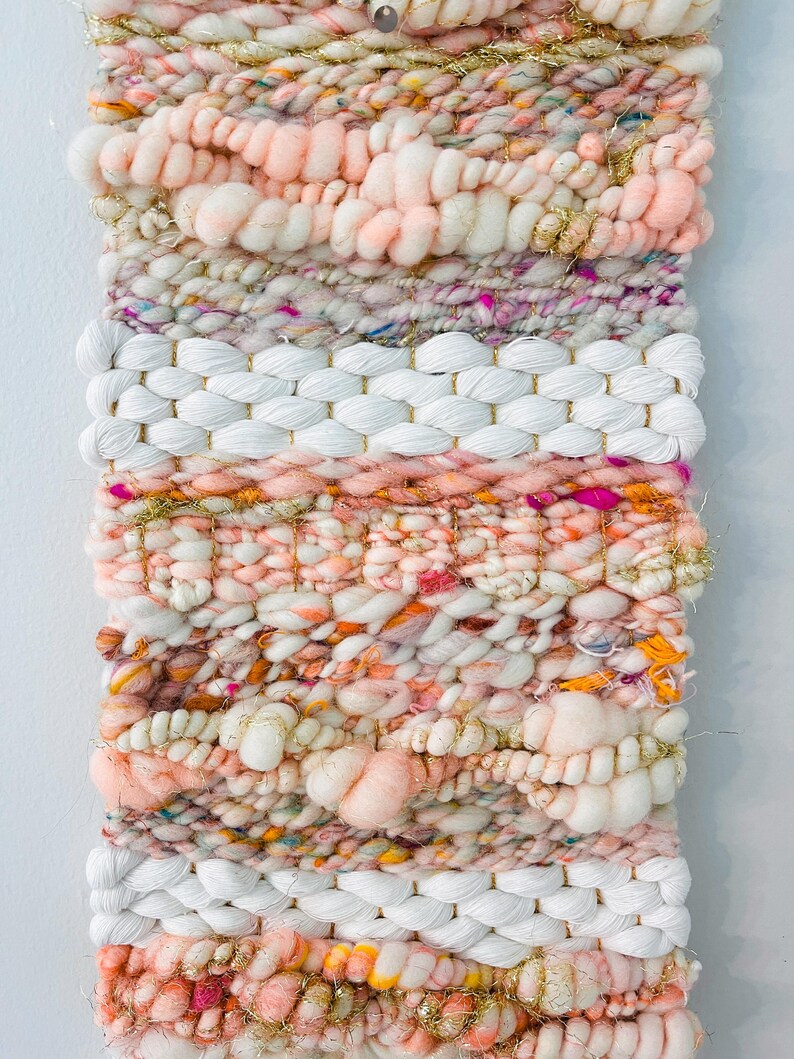 May include: A pink, white, and gold woven wall hanging with a textured, fluffy design. The hanging is made with yarn and has a variety of colors and textures.