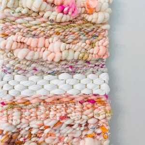 May include: A pink, white, and gold woven wall hanging with a variety of textures and embellishments. The hanging is made with yarn and features a white woven section in the middle.