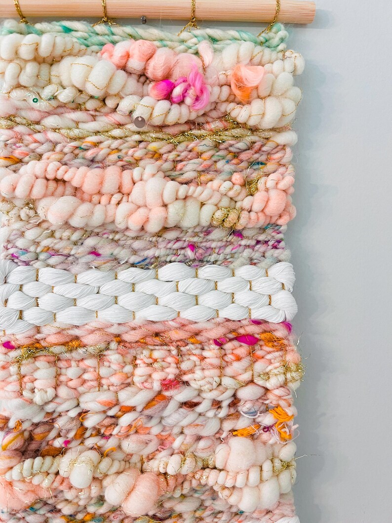 May include: A pink, white, and gold woven wall hanging with a variety of textures and embellishments. The hanging is made with yarn and features a white woven section in the middle.