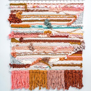 May include: A colorful woven wall hanging with a variety of textures and colors, including pink, orange, brown, and white. The hanging has a fringe border and is made with a variety of yarns.