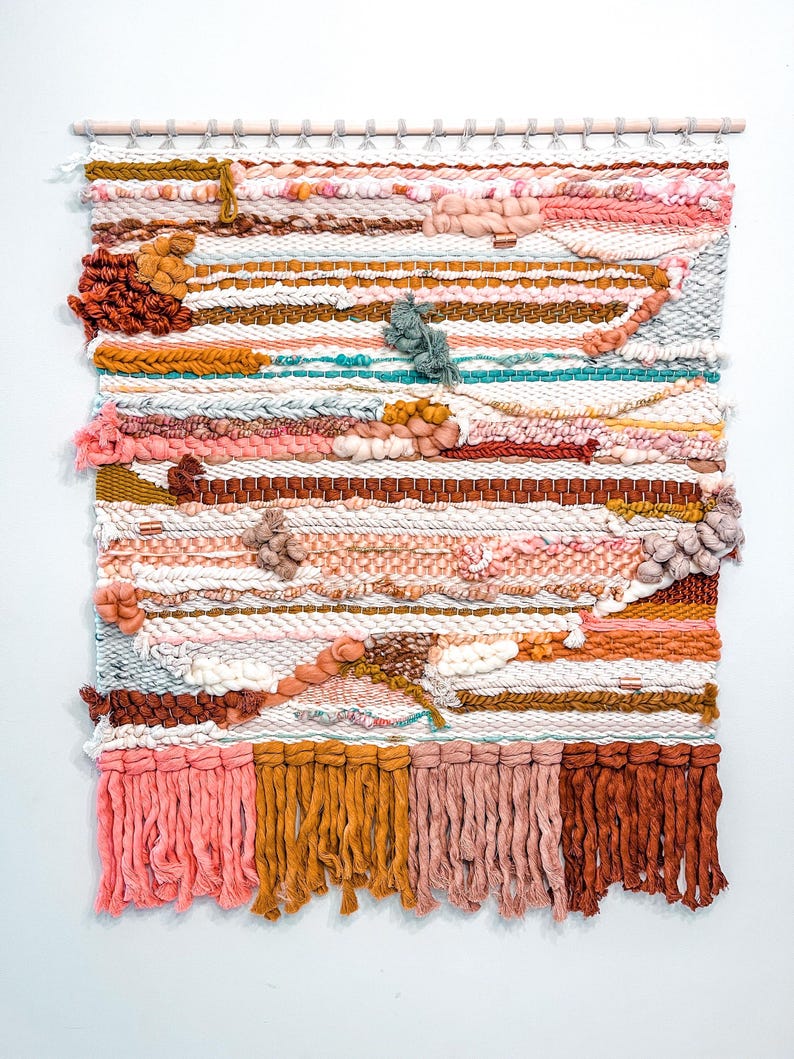 May include: A colorful woven wall hanging with a variety of textures and colors, including pink, orange, brown, and white. The hanging has a fringe border and is made with a variety of yarns.