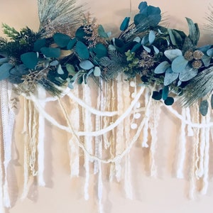 May include: A white and cream garland featuring a mix of textures, including yarn, lace, and pom-poms. The garland is adorned with sprigs of eucalyptus and pine.