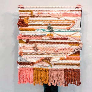 May include: A woven wall hanging with a colorful abstract design. The hanging features a variety of textures and colors, including pink, brown, white, and yellow. The hanging has a fringe border.