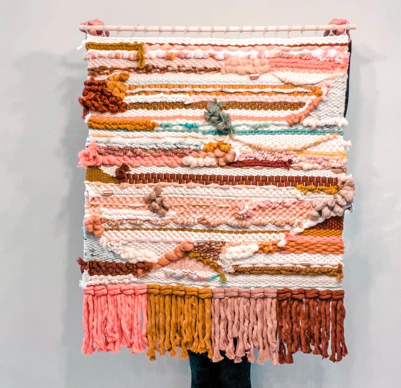 May include: A woven wall hanging with a colorful abstract design. The hanging features a variety of textures and colors, including pink, brown, white, and yellow. The hanging has a fringe border.