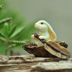 May include: A white rabbit figurine sitting in a small wooden boat with a leaf paddle. The rabbit is holding the paddle with its front paws. The boat is sitting on a piece of driftwood.