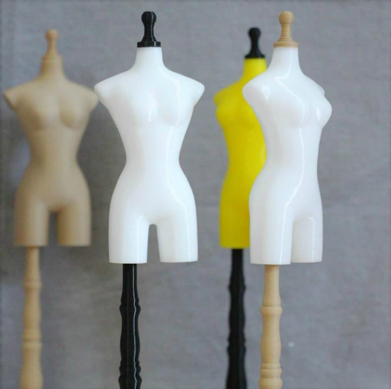 May include: Three different color mannequin torso forms on stands. The mannequins are white, tan, and yellow. The stands are black, tan, and black.