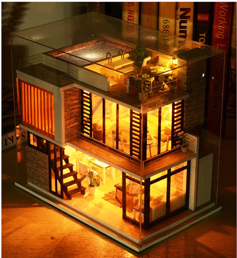 May include: A miniature dollhouse with a glass roof and a lit interior. The dollhouse has two levels, a rooftop pool, and a staircase. The dollhouse is made of wood and has a modern design.