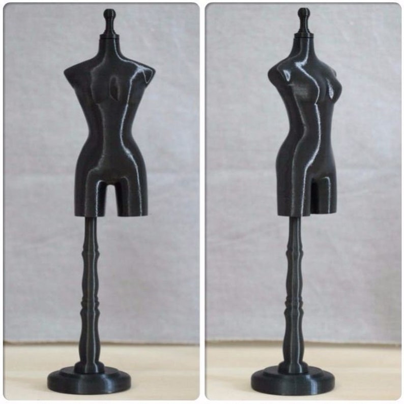 May include: A black mannequin torso on a black stand. The mannequin is made of plastic and has a smooth, glossy finish. The stand has a round base and a fluted column.