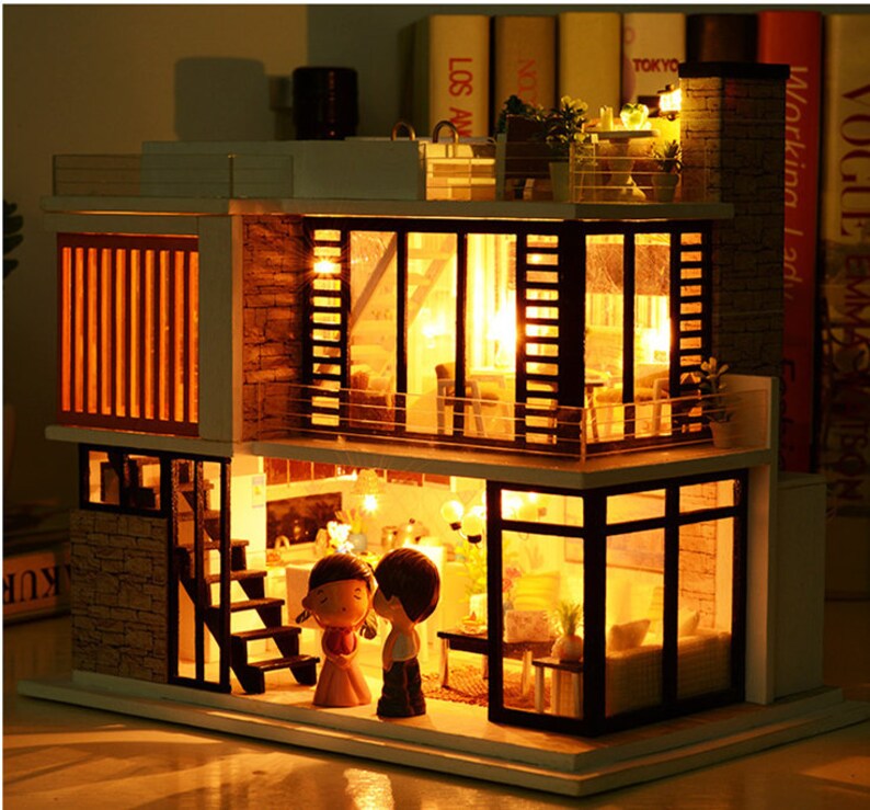 May include: A miniature dollhouse with a white exterior and black windows. The dollhouse has three levels and is lit up with warm white lights. There are two figures standing in front of the dollhouse, a girl with long brown hair and a boy with short brown hair. The dollhouse is on a wooden surface.