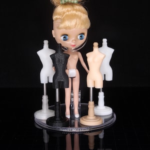 May include: A doll with blonde hair and blue eyes stands in front of four miniature dress forms. The doll is wearing a white skirt and a black and white headset. The dress forms are on stands and are black, white, and beige.