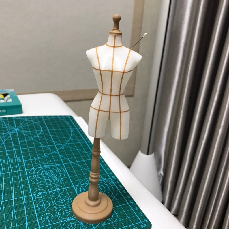 May include: A small white dress form with a grid pattern in brown, standing on a wooden base. The dress form has a pin in the shoulder.
