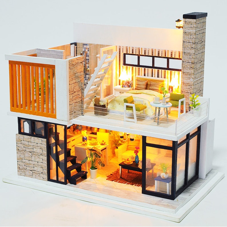 May include: A miniature dollhouse with two levels, featuring a modern interior design. The dollhouse has a white exterior with stone accents and black windows. The interior is furnished with a bedroom, living room, and kitchen, complete with miniature furniture and accessories.