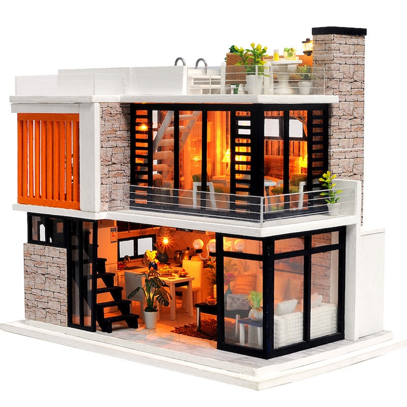May include: A miniature dollhouse model of a modern two-story house with a white exterior, black windows, and a brown brick facade. The house has a balcony on the second floor and a small patio on the first floor. The house is lit up with small lights inside.