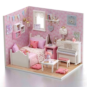 Puede incluir: Un dormitorio de casa de muñecas en miniatura con una cama blanca, ropa de cama rosa, un piano blanco y una mesa pequeña con una taza de té y un platillo. Las paredes son rosas con un patrón floral y hay varios artículos decorativos en toda la habitación.