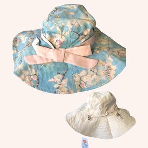 Puede incluir: Dos sombreros para el sol. Uno es azul claro con un estampado floral y un lazo rosa. El otro es un sombrero color crema con ala ancha. Ambos sombreros están diseñados para proteger del sol.