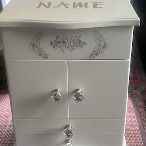 May include: White wooden jewelry box with a floral design and a clear glass knob. The box has three drawers and a hinged lid. The lid has the word "NAME" written on it in gold glitter.