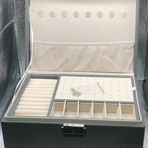 May include: A gray lockable jewelry box with a key, a white interior, and compartments for rings, earrings, and necklaces.