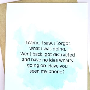 May include: A white greeting card with a humorous quote in black text, set against a light blue watercolor wash. The card is placed on a brown envelope, creating a simple, clean design.