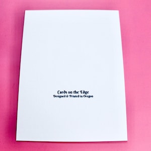 May include: A white greeting card with the text "Cards on the Edge Designed & Printed in Oregon" in dark blue. The card is on a pink background.
