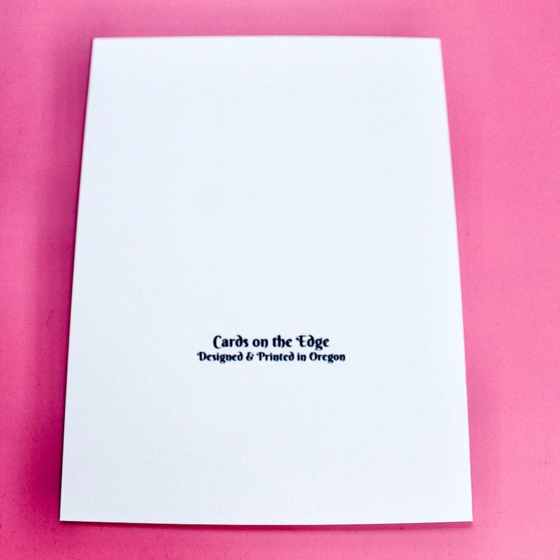 May include: A white greeting card with the text "Cards on the Edge Designed & Printed in Oregon" in dark blue. The card is on a pink background.