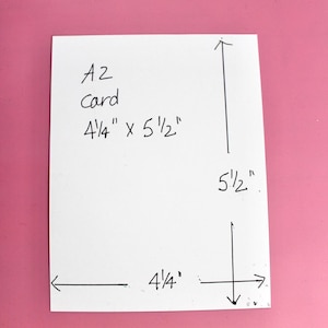May include: A white A2 card with handwritten dimensions of 4 1/4" x 5 1/2". The measurements are indicated with arrows. The card is on a pink background.