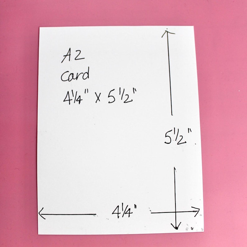 May include: A white A2 card with handwritten dimensions of 4 1/4" x 5 1/2". The measurements are indicated with arrows. The card is on a pink background.