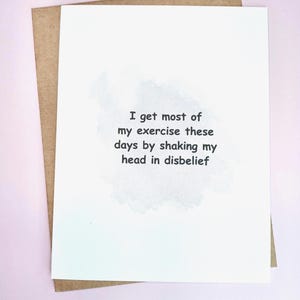 May include: A white greeting card with the text "I get most of my exercise these days by shaking my head in disbelief" in black font. The card is inside a brown envelope, set against a pink background.
