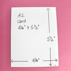 K&ouml;nnte beinhalten: Eine wei&szlig;e A2-Karte mit den Ma&szlig;en 10,8 cm x 14 cm wird vor einem rosa Hintergrund gezeigt. Die Abmessungen der Karte sind deutlich mit schwarzer Tinte beschriftet, einschlie&szlig;lich des Textes "A2 card". Pfeile zeigen die L&auml;nge und Breite an.