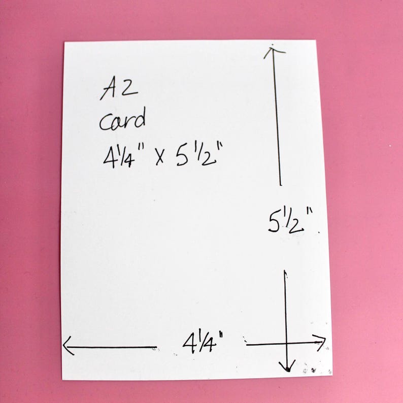 K&ouml;nnte beinhalten: Eine wei&szlig;e A2-Karte mit den Ma&szlig;en 10,8 cm x 14 cm wird vor einem rosa Hintergrund gezeigt. Die Abmessungen der Karte sind deutlich mit schwarzer Tinte beschriftet, einschlie&szlig;lich des Textes "A2 card". Pfeile zeigen die L&auml;nge und Breite an.