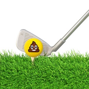 May include: A yellow golf ball with a brown poop emoji on it is being hit by a silver golf club. The ball is sitting on a green grassy surface.