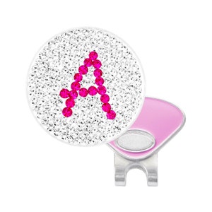 May include: A golf ball marker with a silver base and a pink accent. The round marker is encrusted with clear crystals and features a bright pink letter "A". The marker is designed to attach to a golf hat or visor.