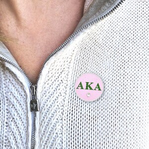 May include: A light pink round button with the letters "AKA" in green glitter. A small, clear gem is centered below the letters. The button is pinned to a white knit sweater.