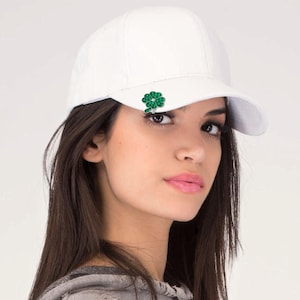 May include: A white baseball cap with a green shamrock emblem on the front. The cap is made of a smooth fabric and has a curved brim. The shamrock is a four-leaf clover design.
