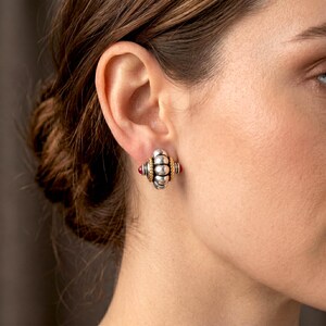 May include: Silver and gold-toned stud earrings with a unique design. The earrings feature a central section with a gold band and two red gemstones. The earrings have a textured, beaded appearance.