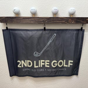 May include: A black banner with white text that reads "2nd Life Golf" and "Giving Old Clubs A Second Chance". A white outline of a golf club is above the text. The banner is hanging from a dark brown wooden shelf with four white golf balls on top.