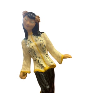 May include: A ceramic figurine of a woman wearing a white and yellow patterned top and black pants. She has black hair and a flower crown. The figurine is standing on a white base.