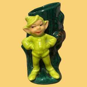 May include: A green ceramic figurine of a whimsical elf standing in a green tree trunk planter. The elf has a pointed hat, large eyes, and a mischievous smile.