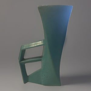 May include: A matte teal ceramic vase with a tall, flared opening and a geometric, angular side handle. The structure features sharp, clean lines and a solid, uniform color throughout, creating a distinct silhouette with a wide top and a narrow base.