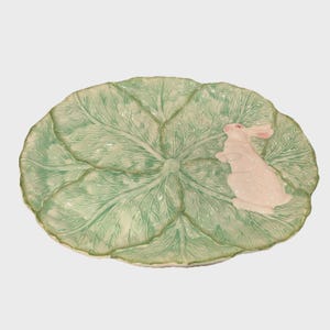 May include: A ceramic serving platter shaped like a large cabbage leaf, in shades of green and white. A white rabbit with pink ears is perched on the leaf. The platter has a textured surface and a scalloped edge.