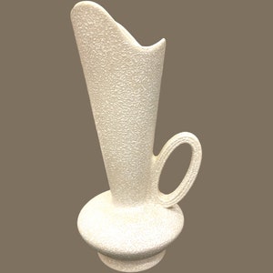 May include: White ceramic vase with a textured surface and a curved handle. The vase has a tall, slender body and a wide, round base.