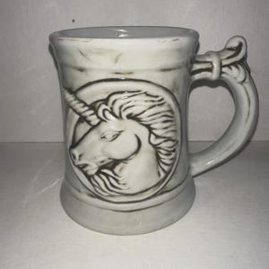 May include: A white ceramic mug with a detailed unicorn head design. The mug has a decorative handle and a slightly textured surface. The unicorn is set in a circular frame. The mug is approximately 5 inches tall.