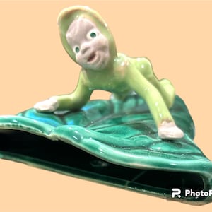 May include: A green ceramic figurine of a pixie or elf with a pointed hat, sitting on a green leaf. The figurine is glazed and has a whimsical expression.