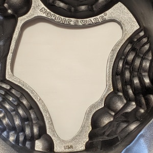May include: A close-up shot of a dark gray, cast aluminum bundt cake pan. The pan has a decorative design with a silver-colored rim and the words "Nordic Ware" and "USA" embossed on it.