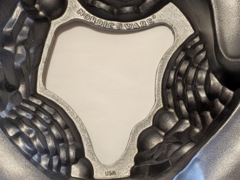 May include: A close-up shot of a dark gray, cast aluminum bundt cake pan. The pan has a decorative design with a silver-colored rim and the words "Nordic Ware" and "USA" embossed on it.